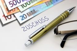 Ein Stapel Geldscheine liegt auf einem Tisch mit dem Schriftzug "Zuschuss". Davor ein Kugelschreiber und eine Lesebrille.
