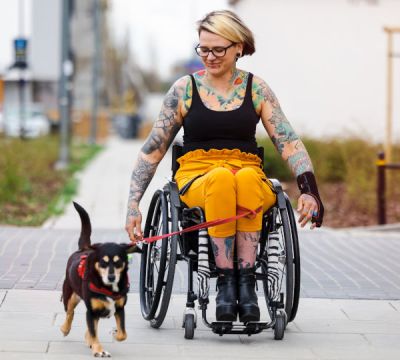 Junge Frau im Rollstuhl fährt mit Ihrem Hund an der Leine spazieren.