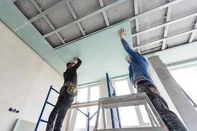 Zwei Handwerker  befestigen Gipskartonplatten an eine mit Blechprofilen abgehängte Zimmerdecke.
