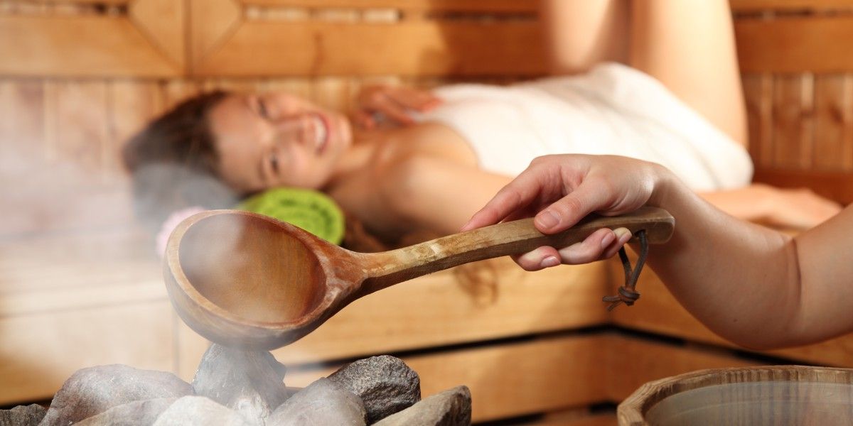 Junge Frau liegt entspannt auf der Bank einer Sauna, während ein Die Steine des Saunaofens mit Wasser übergossen werden.